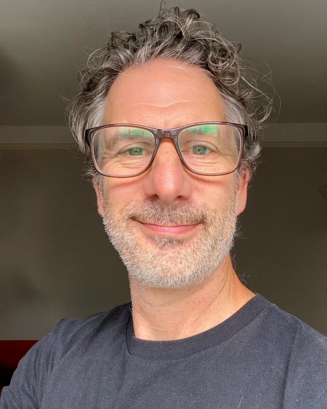 James Willmott, a smiling man with curly grey hair, glasses and a short grey beard, wearing a navy T-shirt